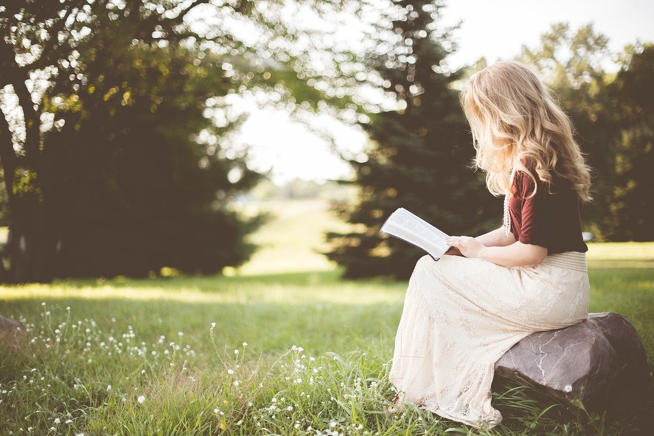 A women sitting on a rock in a serene outdoor setting, engrossed in reading a Bible.